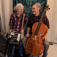 A woman holding a recorder and a man with a cello sitting in front of a small electronic keyboard.